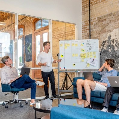 Three men in a room, two sitting and watching one male presenting a board of future plans.